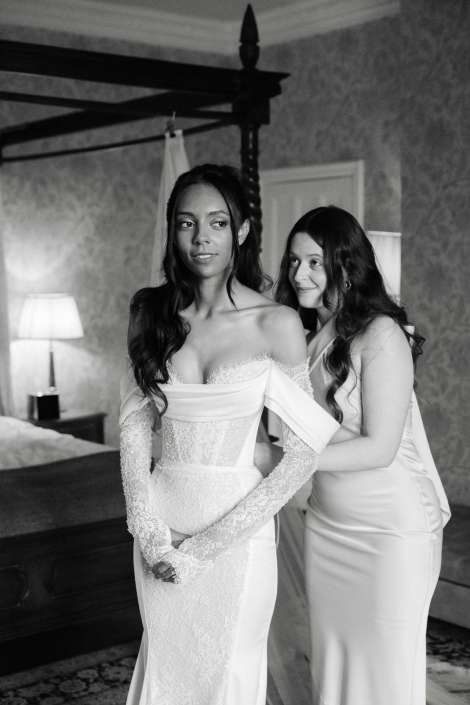 bride getting ready with friend A bride in a lace off-the-shoulder wedding gown stands with her hands clasped as a bridesmaid helps adjust her dress in a bedroom.