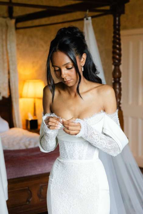 bride adjusting lace wedding dress Bride in a white off-shoulder lace wedding gown adjusting her bodice in a warmly lit bedroom.