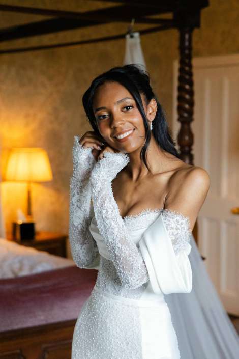 bride in lace wedding dress A smiling bride in a white lace wedding dress with long lace sleeves stands indoors in a warmly lit room.