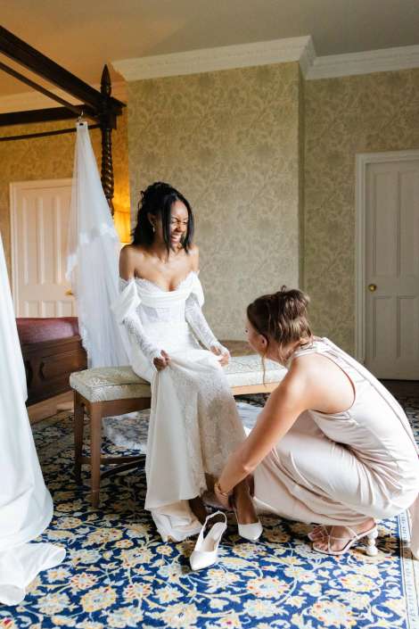 bride’s wedding shoes fitting A bride in a white lace wedding gown sits on a bench as a bridesmaid crouches to help her put on white high-heeled shoes in a room with patterned wallpaper.