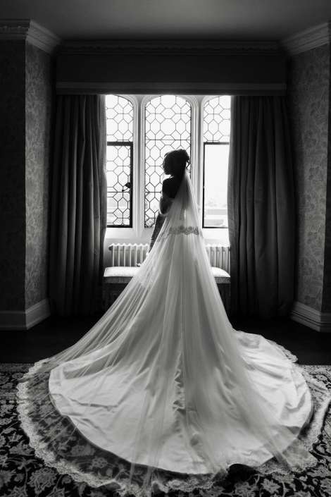 bride in long train gown Back view of a bride in a long wedding gown with a veil, standing by a large arched window.