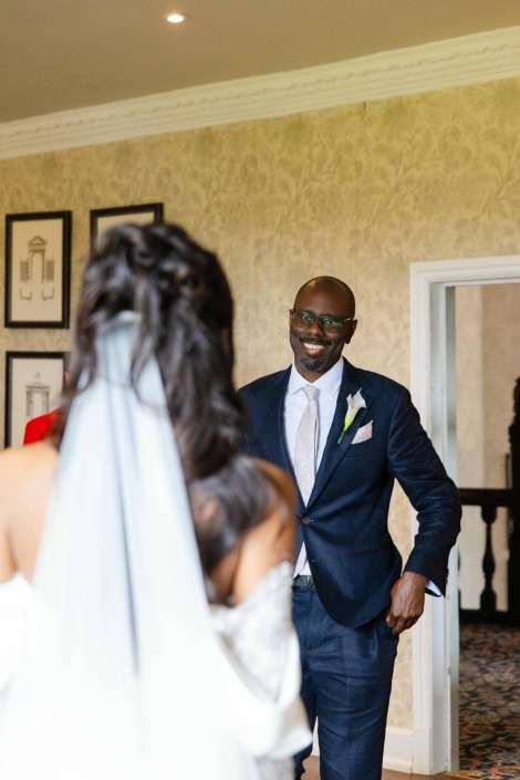 groom smiling at bride A smiling groom in a navy suit with a boutonniere approaches a bride in a white wedding dress indoors.