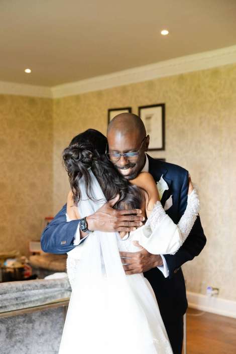 bride and groom embrace A groom in a navy suit and glasses embraces a smiling bride in a white wedding dress with a veil indoors.