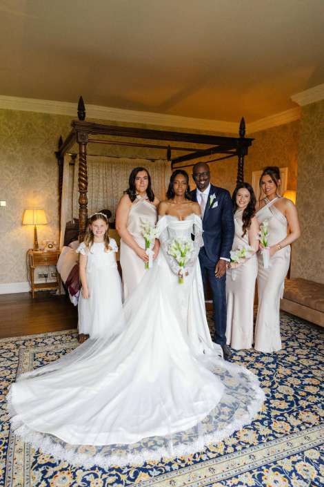 bride and groom with bridesmaids Bride in an off-the-shoulder white wedding gown with a long train stands with bridesmaids in pale pink dresses, a groom in a suit, and a flower girl in white in a luxurious four-poster bedroom.