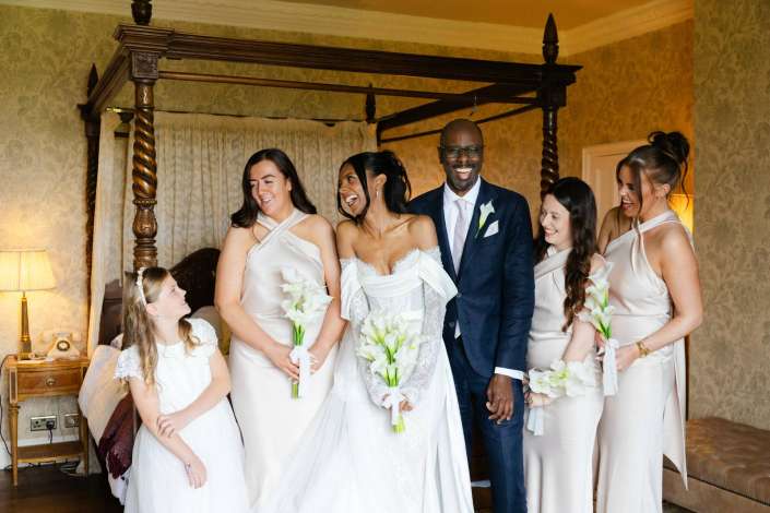 joyful bridal party moment Bride in a white wedding dress with bridesmaids in cream gowns and a smiling groom in a navy suit posing in a vintage bedroom with a four-poster bed.