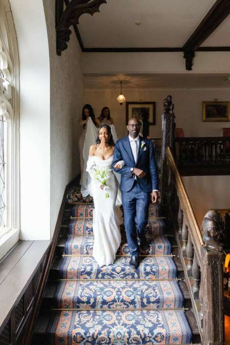 bride and groom descend staircase A bride in a white wedding gown holding a bouquet walks arm-in-arm with a man in a navy suit down a patterned carpeted staircase.