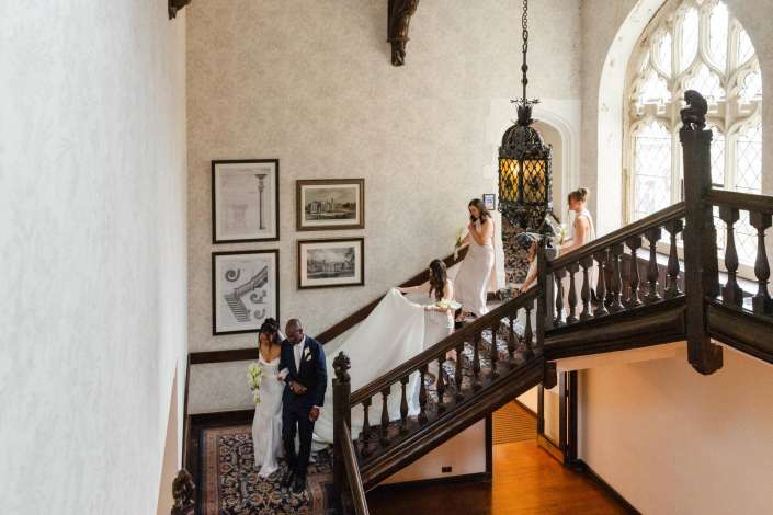 bridal party on grand staircase Bride and groom descend a grand wooden staircase as bridesmaids in white dresses assist, with an ornate hanging lantern and arched windows in a historic mansion.