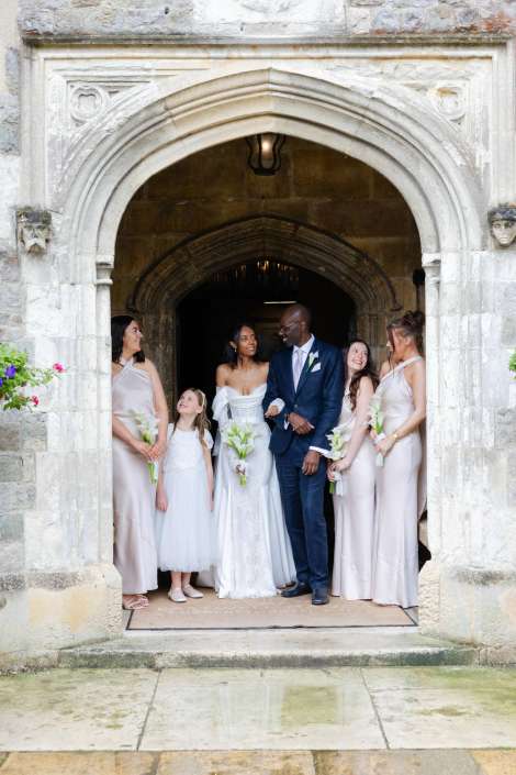 wedding party under stone arch Wedding party posing in a historic stone arch doorway, with the bride in a white gown, the groom in a navy suit, and bridesmaids in pale dresses holding white flowers.