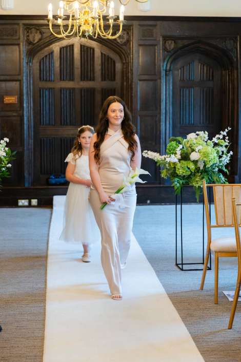 elegant bride-to-be walking down aisle A woman in a pale pink dress walks down a white aisle holding a white lily, followed by a young girl in a white dress in a formal setting.
