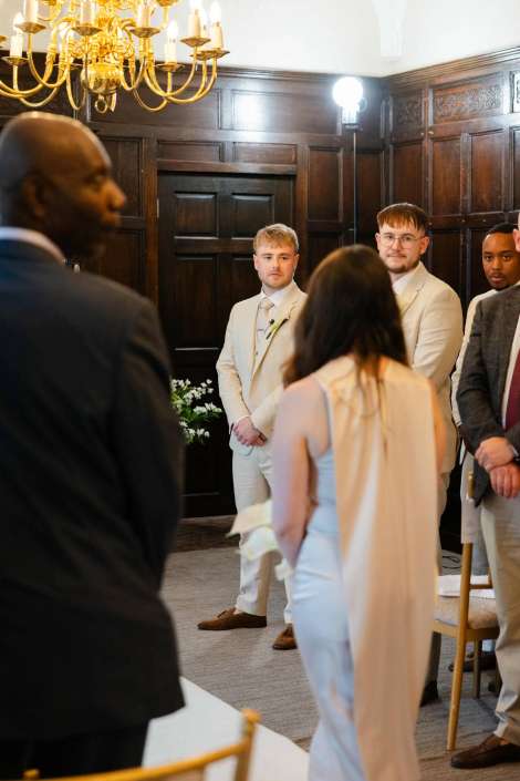 elegant indoor wedding ceremony Several people in light-colored suits stand in a dark wood-paneled room under a chandelier during a formal ceremony.