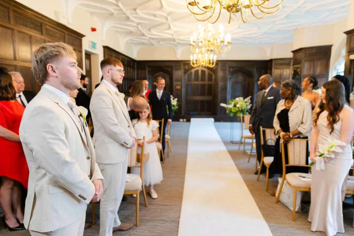 elegant indoor wedding ceremony Guests stand along a white aisle during a formal wedding ceremony in a wood-paneled room with chandeliers.