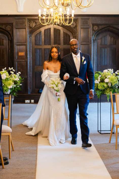 bride walking down aisle Bride in a white gown and groom in a dark suit walk down a white aisle runner at a wedding, with floral arrangements and a chandelier overhead.