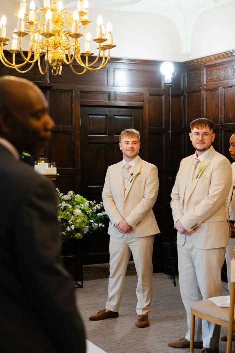 groomsmen in light beige suits Two men in light beige suits stand in a dark wood-paneled room beneath a gold chandelier.