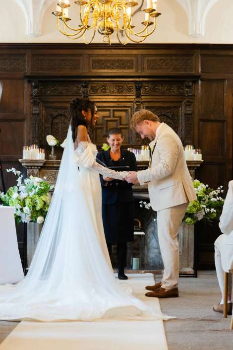 bride and groom ring exchange Bride in a long white wedding gown and veil and groom in a light suit exchange vows in a dark wood-paneled room beneath a chandelier.