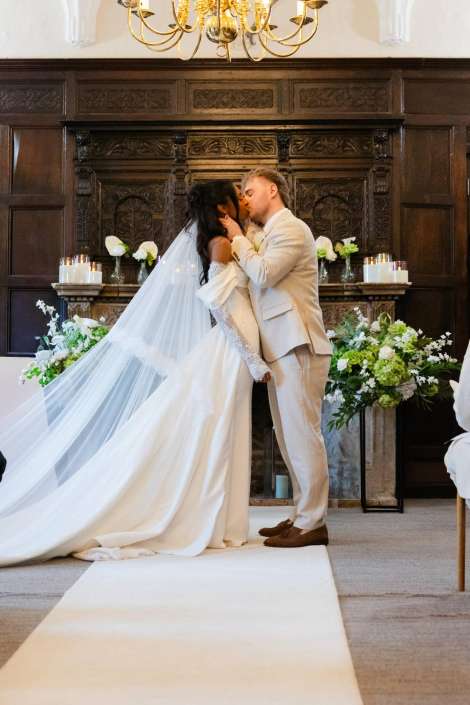bride and groom kiss Bride in a long white wedding gown and veil kisses the groom in a light-colored suit at the altar in a dark wood-paneled chapel with floral arrangements.