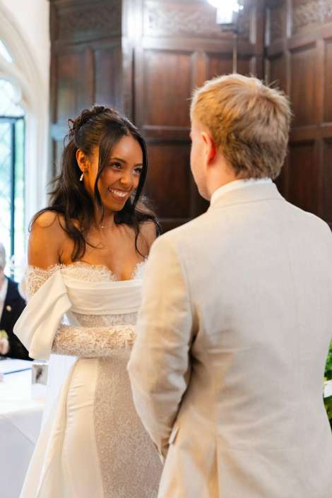 bride smiles at groom A bride in a white off-the-shoulder lace wedding gown smiles at a man in a light suit indoors.