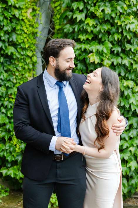 joyful couple amid ivy A bearded man in a suit with a blue tie hugs a smiling woman in a beige dress outdoors against a wall of ivy.