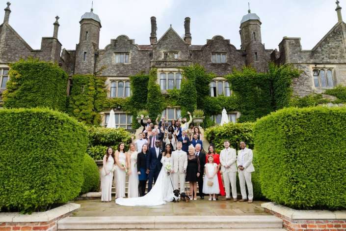 wedding party at ivy covered manor A large wedding party in formal attire poses on stone steps in front of an ivy-covered historic stone building.