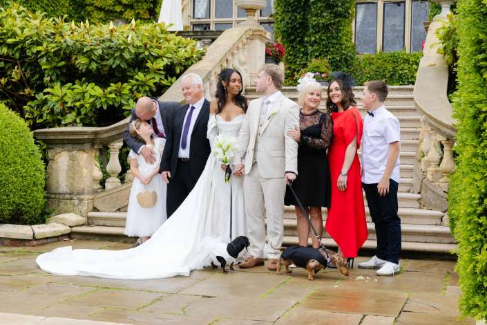 wedding party on stone steps Bride and groom posed with family on a grand stone staircase in a lush garden, with small dogs at their feet.