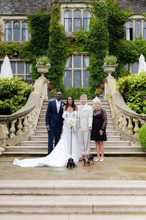 ivy covered mansion wedding party Four people pose on a stone staircase in front of an ivy-covered manor—bride in a white gown, groom in a light suit, a man in a dark suit, and a woman in black—with two small dogs on leashes at their feet.