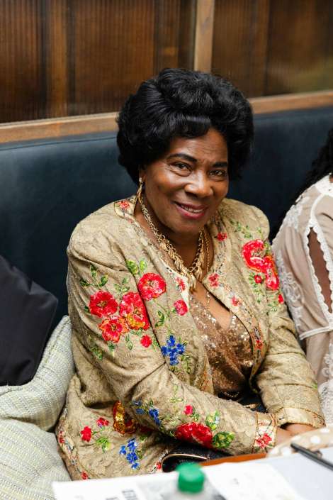 smiling woman in floral embroidered jacket Smiling older Black woman wearing a gold floral-embroidered jacket and a sequined top, with a gold necklace.