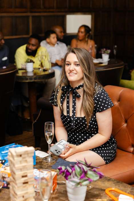 woman in polka dot dress Smiling woman in a black polka-dot dress with a neck bow sits on a brown leather sofa at a social gathering, holding a small grey device with a champagne flute, a wooden block stack, and a small potted plant on the table nearby.