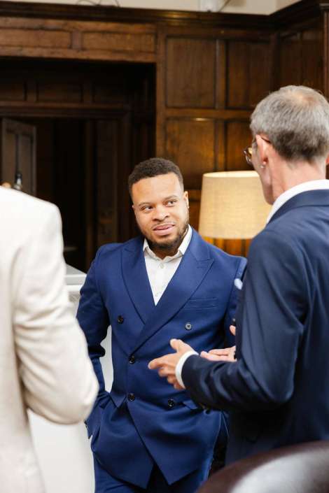 blue double-breasted suit conversation A man in a navy blue double-breasted suit talks with two colleagues in a wood-paneled room.