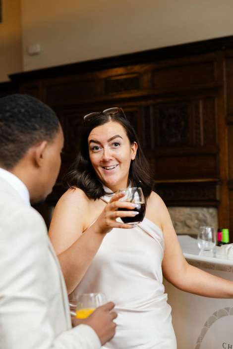woman toasting with red wine A smiling woman in a white dress holds a glass of red wine while chatting with a man in a white suit at a social event.
