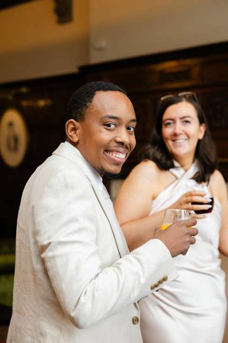 joyful couple at social event Smiling man in a light-colored suit holding a glass, with a woman in a white dress beside him holding a drink in the background.