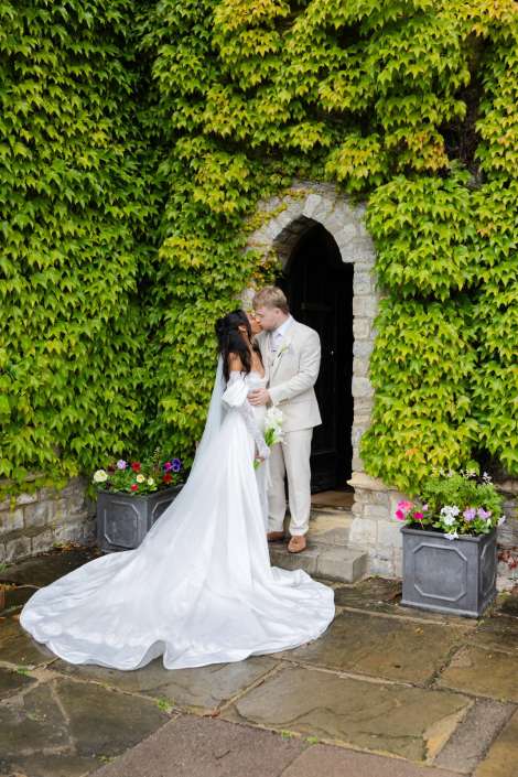 bride and groom kiss under ivy Bride in a long white wedding gown and veil kissing a groom in a light suit in front of a stone arched doorway covered in green ivy.