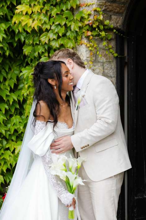 bride and groom embrace A bride in a white lace wedding gown with a veil, holding a bouquet of white calla lilies, smiles as she embraces a groom in a light beige suit beside an ivy-covered stone wall.