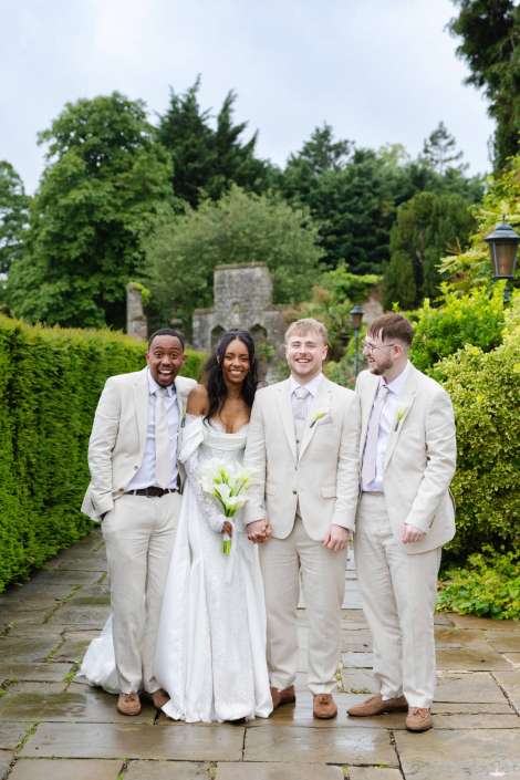 outdoor bridal party portrait Bride in a white wedding gown stands with three groomsmen in light beige suits on a stone path in a green garden, all smiling.