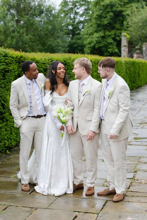 outdoor wedding party portrait Bride in a white lace wedding dress holding a bouquet chats with three groomsmen in beige suits along a stone garden path.