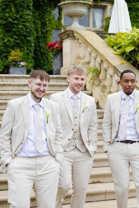 cream suits wedding party Three groomsmen in cream-colored suits pose on an outdoor stone staircase, smiling.