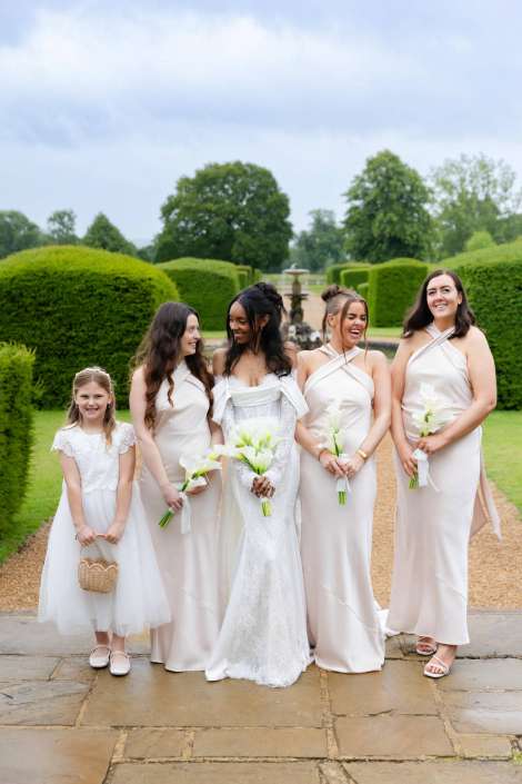 garden wedding party portrait Bride in a white lace gown stands with four bridesmaids in pale blush dresses and a young flower girl in white, all holding white flowers in a formal garden with hedges and a fountain.