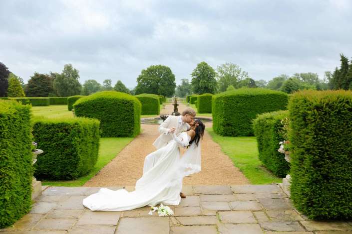 elegant garden wedding portrait Bride and groom kiss as the groom dips the bride in a formal garden with neatly trimmed hedges and a fountain in the background.