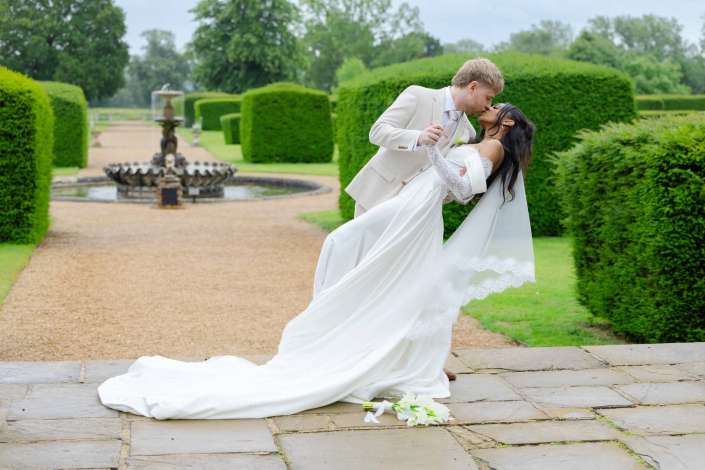 garden wedding kiss A bride and groom kiss as he lifts her in a formal garden with neatly trimmed hedges and a fountain in the background.