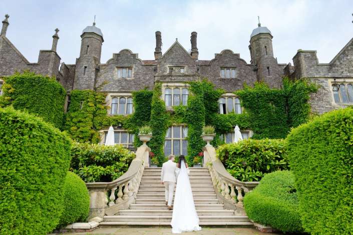 bride and groom on ivy staircase A bride and groom in wedding attire ascend a grand stone staircase toward an ivy-covered, castle-like stone building with towers.