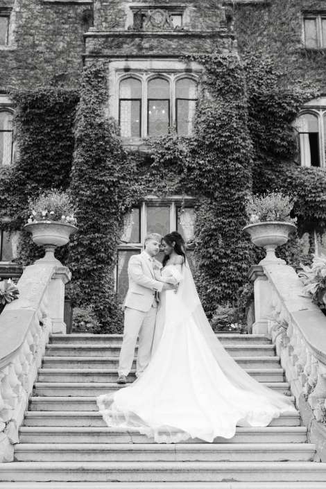 wedding couple on grand staircase Bride and groom embrace on a grand stone staircase before an ivy-covered historic building, the bride in a long flowing wedding gown.