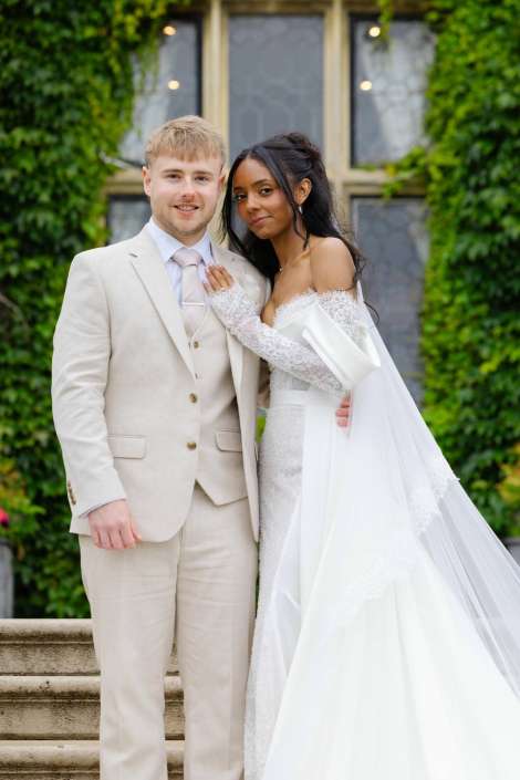 bride and groom portrait Bride and groom pose on stone steps outdoors, the bride in a white lace off-the-shoulder gown with a long veil and the groom in a light beige suit.