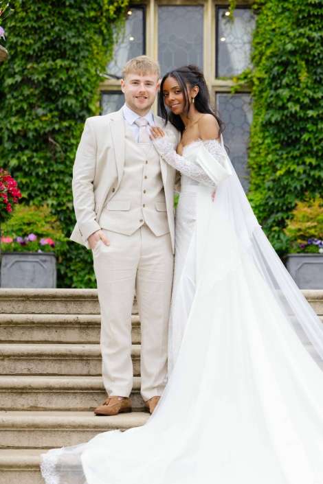 bride and groom on steps Bride in a white wedding gown with a long train stands beside a groom in a beige suit on stone steps, in front of an ivy-covered wall.