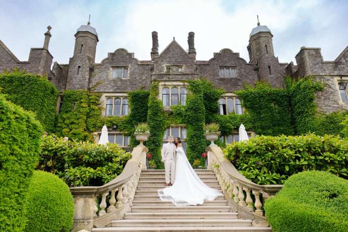 ivied castle wedding staircase Bride and groom in wedding attire stand on a grand stone staircase in front of an ivy-covered castle-like mansion.