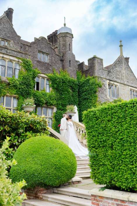 ivy-clad castle wedding kiss Bride and groom kiss on a stone staircase in front of an ivy-covered historic manor with neatly trimmed hedges.