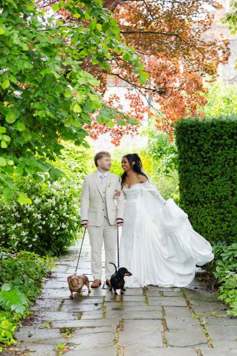 bride and groom walking dogs A bride in a white wedding gown and a groom in a light suit walk two dachshund dogs on leashes along a stone garden path surrounded by lush greenery and autumn leaves.