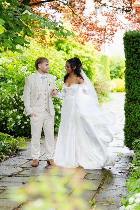 romantic garden wedding walk A bride in a white wedding gown and a groom in a light beige suit walk together along a stone garden path, smiling at each other amid lush greenery and autumn foliage.