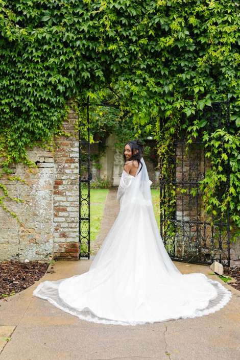 bridal portrait under ivy arch Bride in a flowing white wedding gown with a long train stands at an ivy-covered archway, looking over her shoulder.