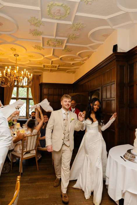 bride and groom entrance A bride in a white wedding gown and a groom in a light beige suit walk hand in hand into a wood-paneled wedding reception, greeted by cheering guests.