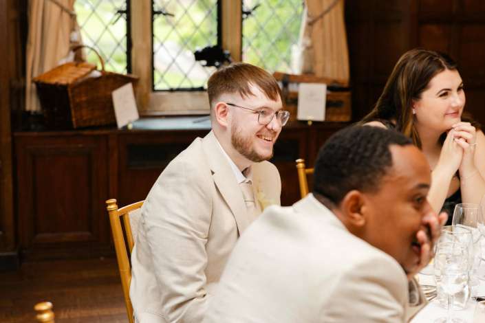 smiling guest at reception A man in a light beige suit and glasses smiles at a dinner table with other guests in a wood-paneled room.