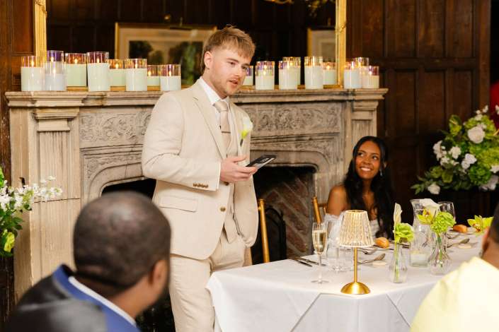 groom delivers wedding toast A man in a light beige suit delivers a speech at a wedding reception beside a candlelit fireplace.
