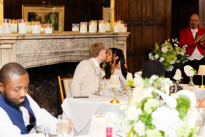 bride and groom kiss at reception A couple kisses at a wedding reception table decorated with candles and flowers, while a server in a red jacket looks on.